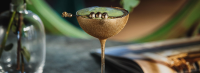 Eleganter Cocktail in einem goldenen Glas auf einem Holzstück, im Hintergrund ein offenes Magazin.
